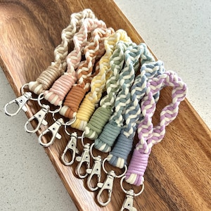 May include: A collection of seven macrame lanyards in various pastel colors, including beige, pink, yellow, green, blue, and purple. Each lanyard features a silver-colored clasp. The lanyards are displayed on a wooden tray.