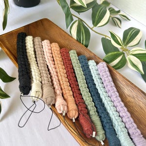 May include: A collection of ten macrame wristlet lanyards in various colours including black, cream, beige, peach, orange, rust, sage green, dark blue, light blue, and lavender, each featuring a black cord loop for attachment to small items.