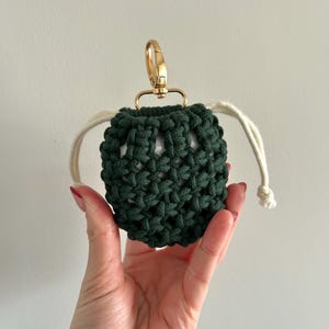 May include: A dark green macrame pouch with a gold-colored clasp and cream-colored drawstring. The pouch is designed to hold small items and is held in a hand.