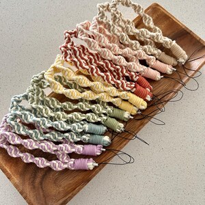 May include: A collection of colorful macrame wrist straps, each with a loop and a small cord for attaching to a phone or other device. The straps are in various colors, including pink, yellow, green, and purple, and are displayed on a wooden tray.