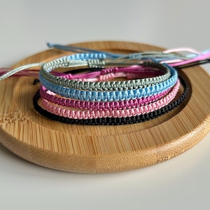 May include: A selection of macrame bracelets in shades of blue, pink, and black, presented on a round wooden tray. The bracelets feature intricate woven patterns, highlighting the craftsmanship. The bracelets are made of woven cord.
