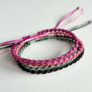 May include: Four macrame bracelets in pink, grey, and black. The bracelets are made of woven cord with a textured, wavy design. The bracelets are stacked, with the black bracelet at the bottom. The bracelets are suitable for everyday wear.