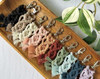 May include: A collection of ten macrame keychains in various colors including black, beige, cream, pink, peach, rust, sage green, light blue, dark blue, and lavender. Each keychain features a knotted cord design and a silver metal lobster clasp.