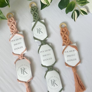 May include: Three macrame Polaroid photo hangers in dusty pink, sage green, and terracotta. Each features a wooden ring at the top, knotted cord sections, and a tassel at the bottom. The hangers display white cards with the text Polaroid Photo Hanger and the Penguin Knots PK logo.