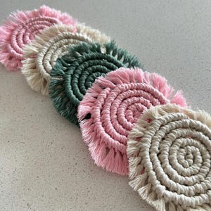 May include: Five round crocheted coasters with fringe. The coasters are in shades of pink, beige, and green.