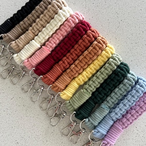 May include: A selection of macrame lanyards in a spectrum of colours, such as black, beige, pink, red, orange, yellow, green, blue, and purple. Each lanyard has a woven design and a silver-coloured metal clasp.