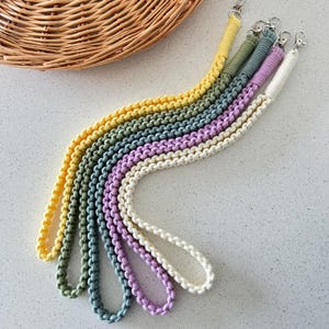 May include: A selection of macrame lanyards in various colours, including yellow, green, blue, purple, and white. Each lanyard has a woven pattern and a silver clasp. The lanyards are displayed on a light surface, with a woven basket visible in the background.