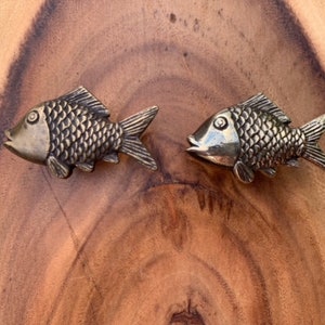 May include: Two silver fish-shaped drawer pulls with detailed scales. The pulls are on a wooden surface.