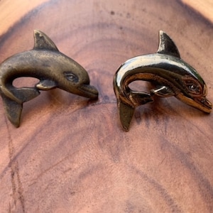 May include: Two gold-toned dolphin figurines, one with a more polished finish and the other with a more matte finish, are displayed on a wooden surface.