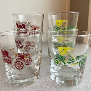 May include: Four vintage glass tumblers with colorful illustrations of antique cars. The tumblers feature red, yellow, and green cars with gold accents. The text on the tumblers reads "Cadillac Model M" and "1909 Packard Roadster".