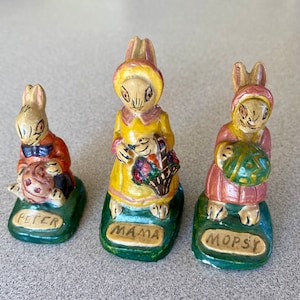 May include: Three ceramic figurines of Peter Rabbit characters. The figurines are painted in yellow, orange, pink, and green. The figurines are standing on green bases with the names "Peter", "Mama", and "Mopsy" written on them.
