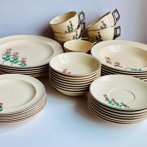 May include: A vintage cream-colored dinnerware set with a floral design. The set includes plates, bowls, saucers, and cups with gold trim. The floral pattern features purple and green accents. A complete set for serving meals.