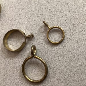 May include: A collection of brass-toned metal curtain rings, including one wider band ring with a small hook attachment and two thinner circular rings, each featuring a small eyelet loop at the top for hanging purposes on a rod.