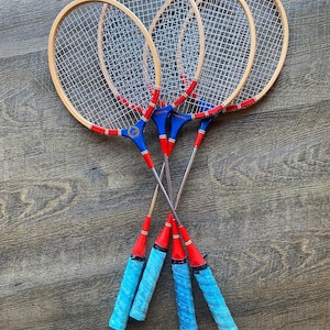 May include: Four vintage badminton rackets with wooden frames and white netting. The handles are wrapped in blue grip tape, and the stems are red and blue. The rackets are arranged on a wooden surface.