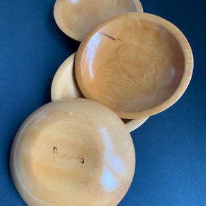 May include: Four wooden bowls with a light, natural finish are arranged on a dark blue surface. The bowls are round with a shallow depth. The bottom of one bowl has the word "flurusing" etched into the wood.