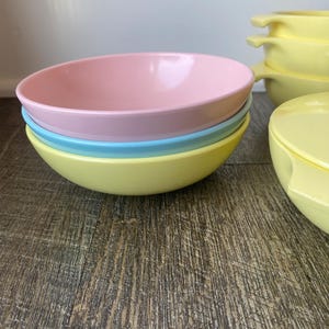 May include: A stack of pastel-colored plastic bowls in pink, blue, and yellow. The bowls have a smooth, rounded design. Additional yellow bowls with handles are visible in the background. The bowls are on a wooden surface.
