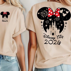 May include: Beige t-shirt featuring a Minnie Mouse design with a red bow and the name "Amelia" on the front. The back of the shirt has a larger Minnie Mouse design with a castle, fireworks, and the text "Disney Trip 2026".