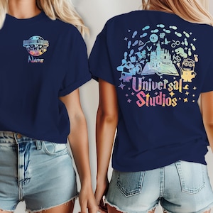 Może przedstawiać: Granatowa koszulka z kolorowym logo Universal Studios z przodu i z tyłu. Logo z przodu zawiera tekst "Name". Z tyłu znajduje się zamek, postacie i napis "Universal Studios".