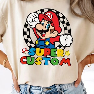 Könnte beinhalten: Ein beiges Kurzarm-T-Shirt mit einer Grafik von Mario aus Super Mario Bros. in einem schwarz-weiß karierten Kreis. Unter der Figur steht der Text SUPER CUSTOM in bunten Blockbuchstaben, begleitet von einem kleinen Pilz und einem Stern.