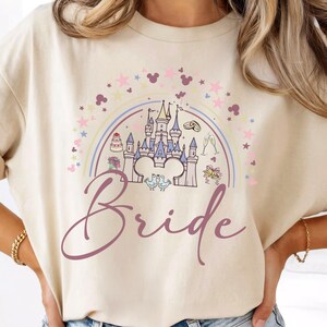 Puede incluir: Camiseta beige con un gráfico colorido que presenta un castillo, un arcoíris y la palabra "Bride" en una fuente cursiva. El diseño incluye estrellas, siluetas de Mickey Mouse y elementos temáticos de boda.