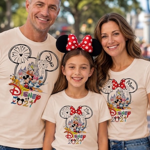 Puede incluir: Tres camisetas de color crema con un gráfico de una silueta de orejas de Mickey Mouse relleno con un castillo y personajes de Disney, fuegos artificiales, el texto Disney 2021, etiquetas individuales para Dad, Mom y Lizy, y una diadema de lunares rojos.
