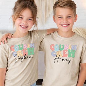 May include: Two beige short-sleeve t-shirts featuring the text "COUSIN CREW" in a multi-coloured, wavy font. Below the text, one shirt displays the name "Sophia" and the other displays the name "Henry" in a black, cursive script font.