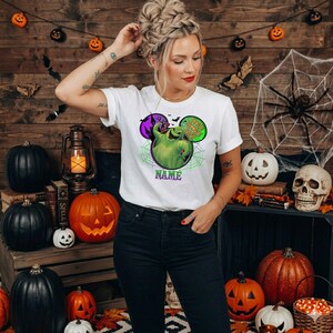 May include: A white short-sleeve t-shirt featuring a graphic of a green Oogie Boogie character inside a Mickey Mouse-shaped outline with spiderwebs, a purple haunted house, and the text "Oogie Boogie Bash" and "NAME" in purple and green.