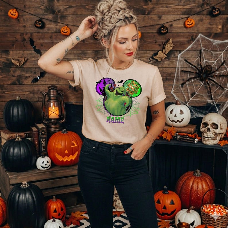 May include: A beige short-sleeve t-shirt featuring a graphic of a green character inside a circular frame with spiderwebs, purple castle silhouettes, and the text "Oogie Boogie Bash" and "NAME" printed in purple and green lettering.