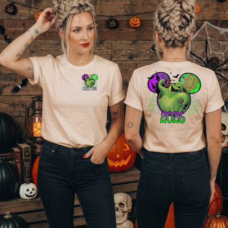 Puede incluir: Una camiseta beige de manga corta con un peque&ntilde;o gr&aacute;fico en el pecho izquierdo que muestra un personaje verde y la palabra CUSTOM. La parte posterior presenta un gr&aacute;fico grande de un personaje verde, una casa embrujada y el texto OOGIE BOOGIE BASH 2026.