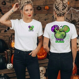Puede incluir: Una camiseta blanca de manga corta con un peque&ntilde;o gr&aacute;fico de un personaje verde en el pecho izquierdo y un gr&aacute;fico grande en la espalda. El dise&ntilde;o trasero muestra un personaje verde, un castillo morado y el texto OOGIE BOOGIE BASH 2026.