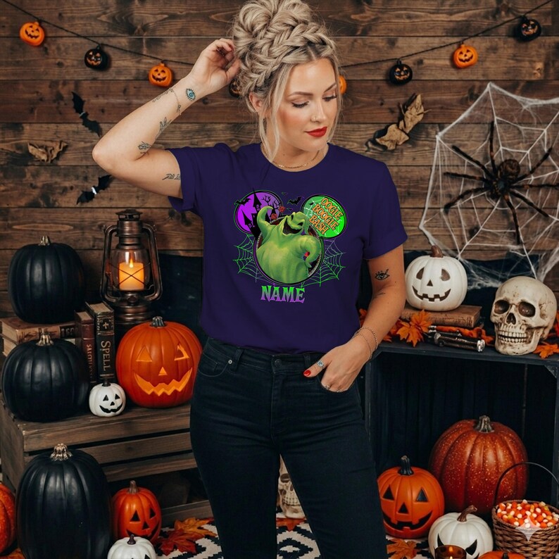 May include: A dark purple short-sleeve t-shirt featuring a graphic of the green Oogie Boogie character from The Nightmare Before Christmas, surrounded by a spiderweb design, a purple castle, and the text Oogie Boogie Bash and NAME.