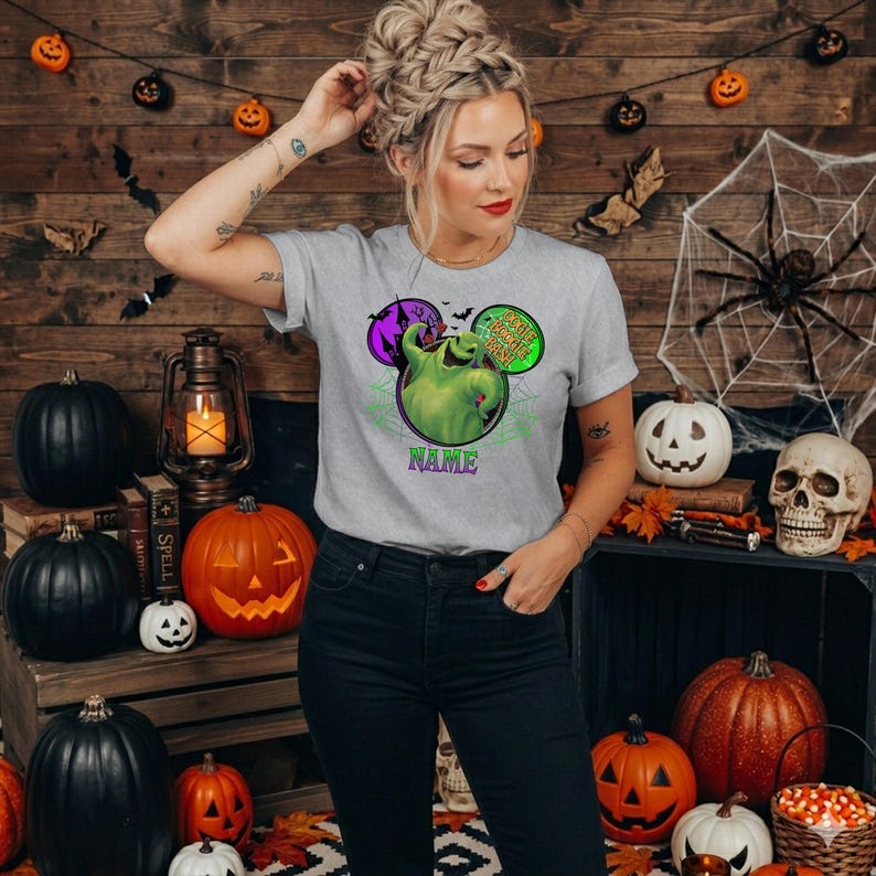 May include: A light gray short-sleeve t-shirt featuring a graphic of a green character from The Nightmare Before Christmas inside a purple and green circular frame with the text Oogie Boogie Bash and the word NAME printed in purple below.