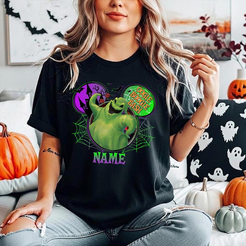 May include: A black t-shirt featuring a graphic of the character Oogie Boogie in a green hue, surrounded by a spiderweb design and mouse-ear shapes. The text OOGIE BOOGIE BASH is printed in green, and the word NAME is printed in purple below the graphic.