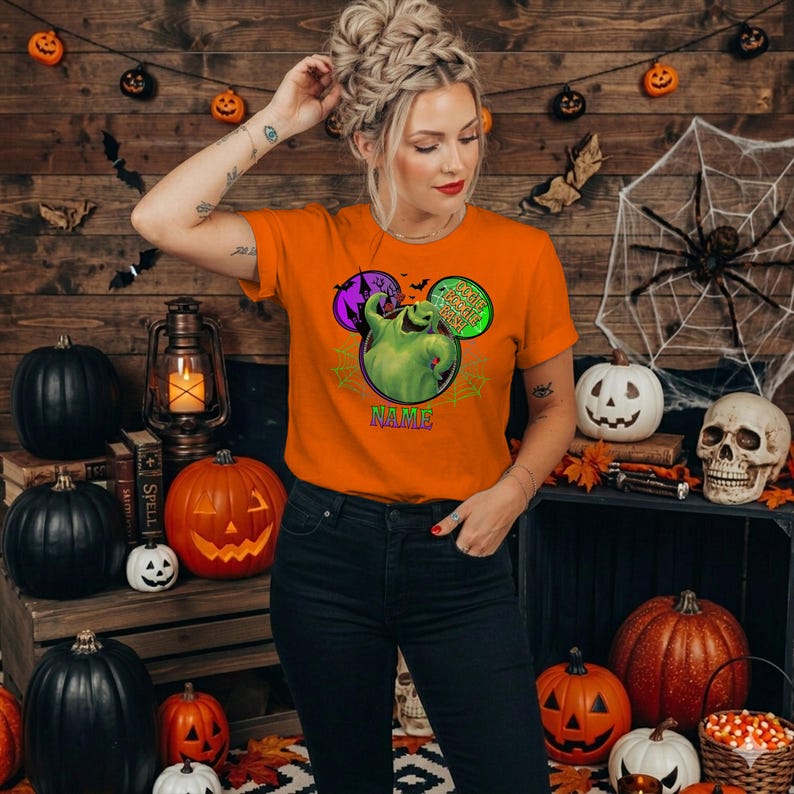 May include: An orange short-sleeve t-shirt featuring a graphic of the green Oogie Boogie character from The Nightmare Before Christmas, surrounded by a purple castle, spiderwebs, and the text Oogie Boogie Man with a space for a name.