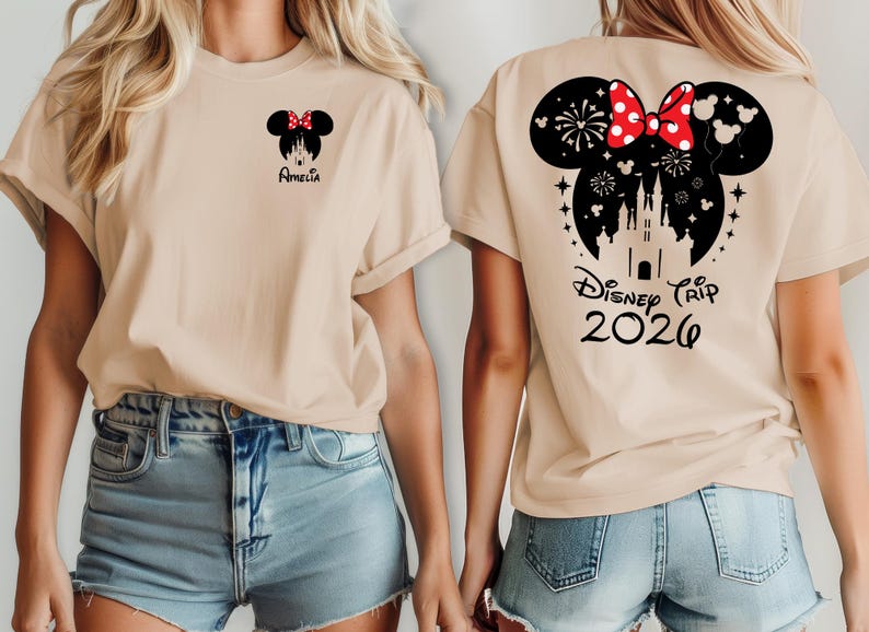 May include: Beige t-shirt featuring a Minnie Mouse design with a red bow and the name "Amelia" on the front. The back of the shirt has a larger Minnie Mouse design with a castle, fireworks, and the text "Disney Trip 2026".