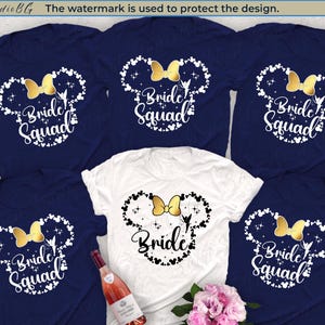 Può includere: Sei magliette blu navy con un contorno bianco delle orecchie di Topolino e il testo "Bride Squad" in bianco con un fiocco dorato. Una maglietta bianca con un contorno nero delle orecchie di Topolino e il testo "Bride" in nero con un fiocco dorato.