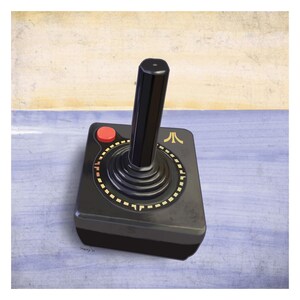Peut inclure: Une manette Atari noire avec un bouton rouge et des détails dorés. La manette a une longue poignée noire et une base carrée avec un design circulaire. Le logo Atari est visible sur la base.