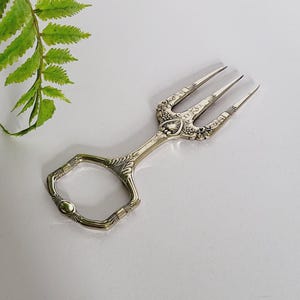 Puede incluir: Pinzas de servir plateadas con un mango decorativo y tres púas. Las pinzas tienen un mango en forma de tijera y están sobre un fondo blanco. Se ve una planta verde en una maceta estampada. Para servir pasteles o repostería.