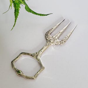 Puede incluir: Tenedor de servir plateado con detalles florales ornamentados. El mango presenta un pequeño acento verde redondo. El tenedor de tres puntas está diseñado para servir pasteles u otros artículos pequeños. Una pieza decorativa para una fiesta de té.