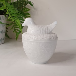 以下が含まれることがあります： 白いセラミック製のアヒルの形をした小物入れで、詳細な蓋が付いています。白い表面に置かれています。箱はテクスチャード加工のデザインです。背景には鉢植えのシダがあります。