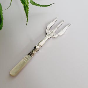 Puede incluir: Un tenedor plateado con tres púas, con detalles ornamentales en la cabeza y un mango con incrustaciones de nácar. El tenedor mide aproximadamente 15 cm de largo, ideal para servir aperitivos o postres. El diseño es elegante y vintage.