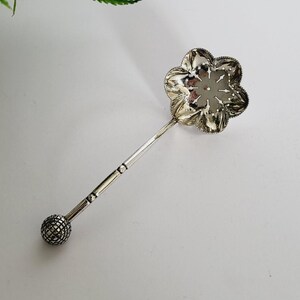 以下が含まれることがあります： 花型のストレーナーと長い柄が付いた銀色のティーインフューザー。ハンドルは、端にテクスチャード加工されたボールが付いています。インフューザーは、左上隅に緑の葉がある白い背景に配置されています。
