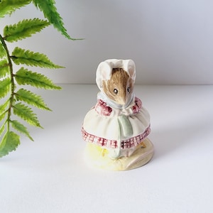 May include: A ceramic mouse figurine, wearing a bonnet and dress. The mouse has brown fur and a white dress with a red and green pattern. The figurine is on a white surface.