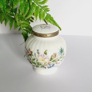 May include: A white ceramic jar with a lid, decorated with a floral pattern in pastel shades. The jar has a ribbed texture and a gold-coloured rim. A green plant in a white and green patterned pot is in the background.