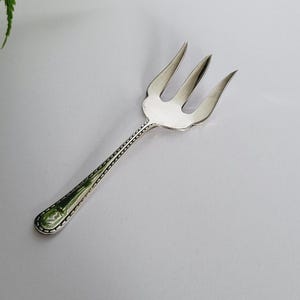 Puede incluir: Un tenedor plateado con tres púas, con un mango detallado con un patrón decorativo. El tenedor se encuentra sobre un fondo gris claro, con una ramita de follaje verde en la esquina superior izquierda.