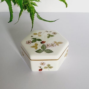 May include: A cream-coloured, hexagonal trinket box with a lid decorated with a strawberry and floral pattern. The box has a gold trim. A green plant in a patterned pot is in the background. The box is set against a white background.