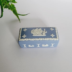 Puede incluir: Pequeña caja de baratijas rectangular azul con detalles en relieve blancos. La caja presenta un diseño clásico con figuras y detalles florales. La caja mide aproximadamente 10 cm de largo y 5 cm de ancho, con tapa.