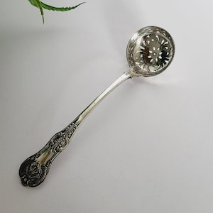 以下が含まれることがあります： ボウルとハンドルに花の模様が入った銀色のレードル。ハンドルには、装飾的で詳細なパターンがあります。レードルは、左上隅に緑のシダの葉がある白い背景に設定されています。