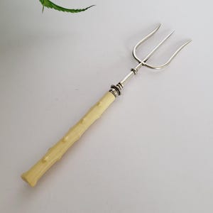 Puede incluir: Un tenedor de servir plateado y color marfil con tres púas. El mango tiene un diseño texturizado en forma de rama. El tenedor está sobre un fondo blanco con una planta verde en la esquina superior izquierda.