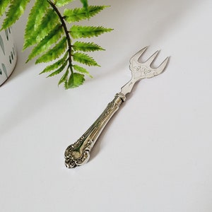 Puede incluir: Un tenedor de servir plateado con tres púas y detalles ornamentales en el mango. El tenedor está sobre una superficie blanca, con una planta de helecho verde en una maceta blanca y azul estampada en el fondo.